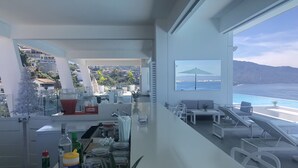 A modern living room with a large window overlooking a pool and ocean. There is a dining area with a bar and various kitchen items visible. - Orchid Condo (Puerto Vallarta)