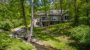 A two-story house with a deck, surrounded by lush greenery and a small stream.