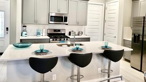 A modern kitchen with grey cabinets, a stainless steel microwave, a stove, and a refrigerator. There are three black bar stools and a large island with blue glassware. - LUXURY Modern Reunion Resort Home . Golf Course Views, Pool Access, near Disney! (Kissimmee)