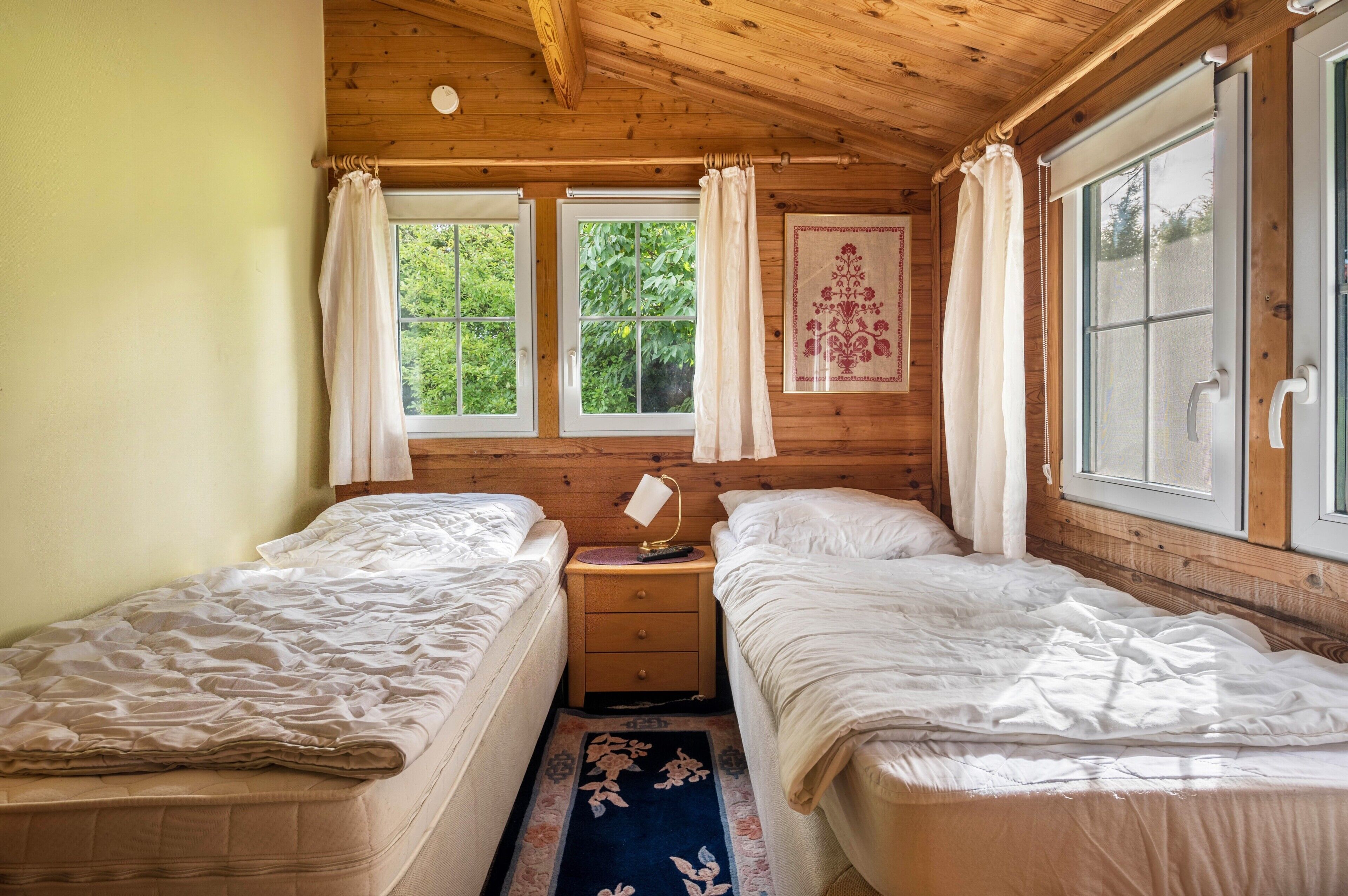 Une chambre dans un chalet en bois, avec deux lits, une table de chevet, une fenêtre avec des rideaux et une porte dotée d’une poignée.