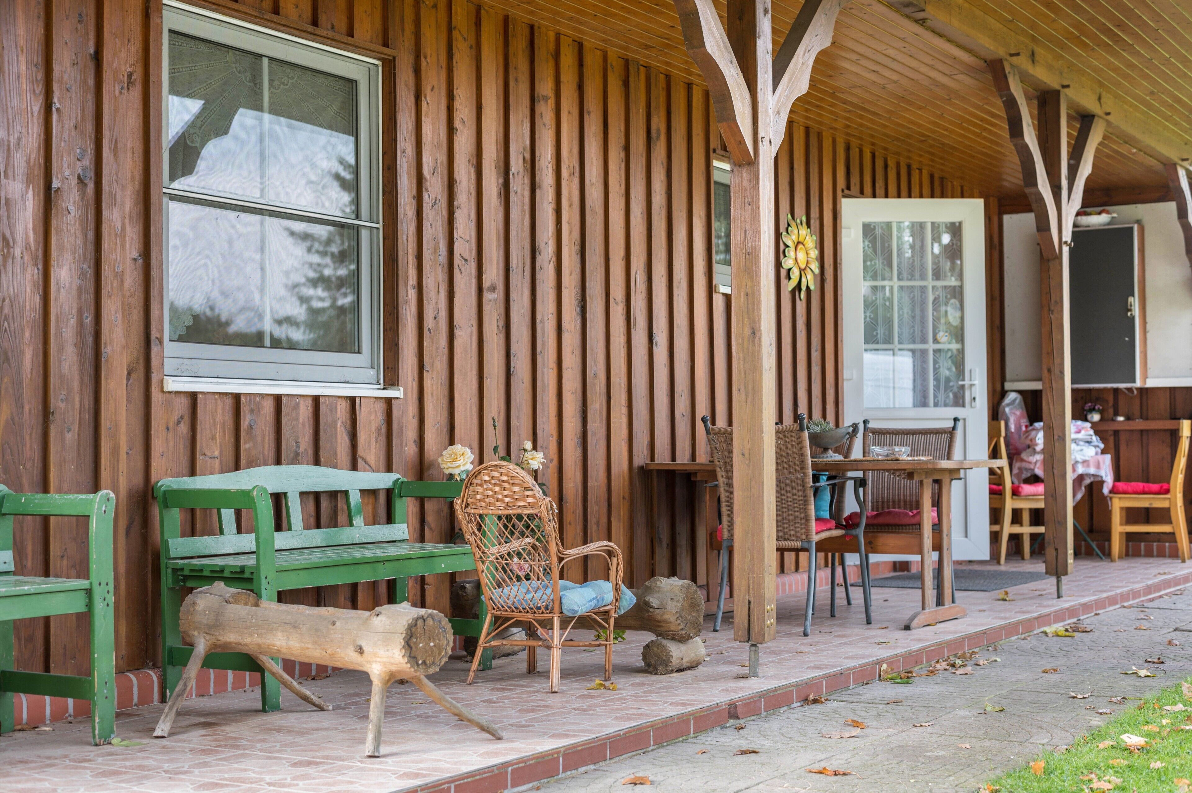 A wooden cabin with a porch, green benches, wicker chairs, and a table with chairs.