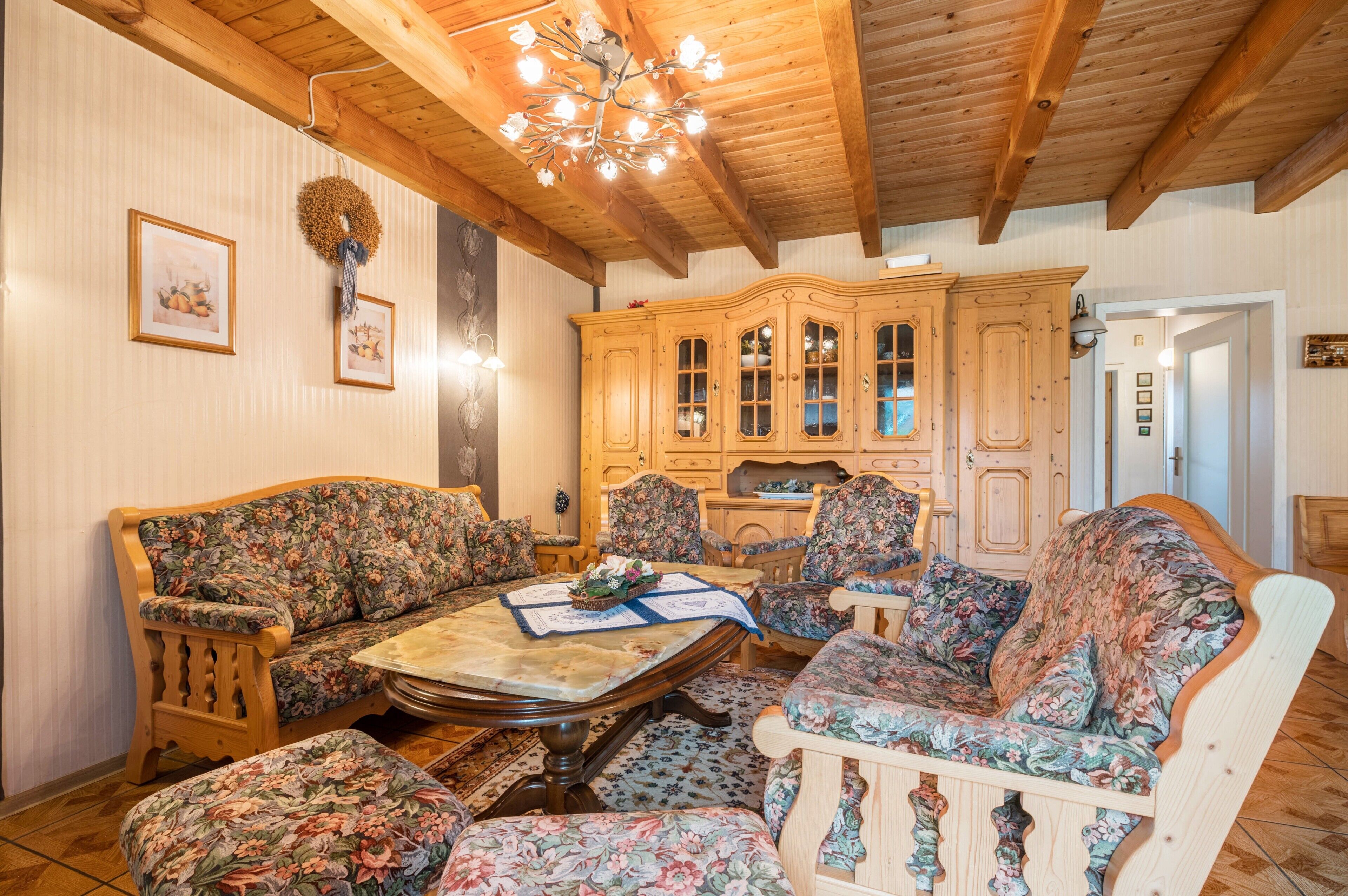 Une pièce avec des poutres apparentes en bois au plafond, un ensemble de meubles de salon à motifs floraux, un buffet en bois et un lustre.
