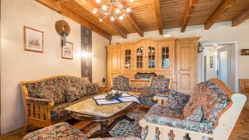 Une pièce avec des poutres apparentes en bois au plafond, un ensemble de meubles de salon à motifs floraux, un buffet en bois et un lustre.