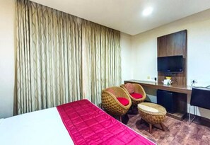 Habitación de hotel con cama, dos sillas de mimbre con cojines rojos, un pequeño reposapiés, un escritorio con televisor de pantalla plana y un armario de madera.