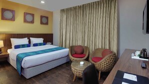 Quarto de hotel com uma cama, duas cadeiras de vime, uma mesa pequena e uma escrivaninha com um controle remoto.