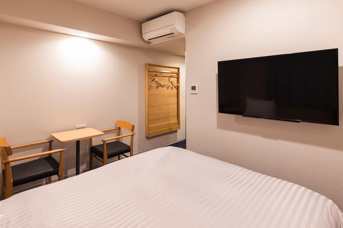 A hotel room with a bed, a TV mounted on the wall, a small table with two chairs, and a wardrobe with a mirror.