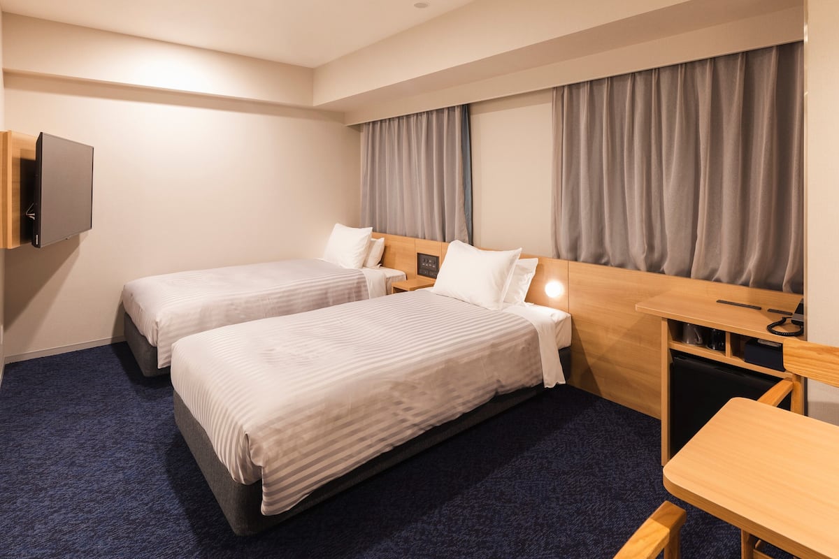A hotel room with two beds, a desk, a television, and a window with curtains.