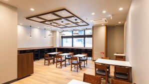 Un interior de restaurante moderno con mesas y sillas de madera, iluminación empotrada en el techo y grandes ventanales que permiten la entrada de luz natural.