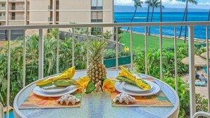 A balcony with a table set for two, overlooking a tropical landscape with palm trees and a view of the ocean. - Oceanview Soothing Waves Kaanapali West MAUI STUDIO (Lahaina)