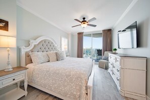 Une chambre avec un grand lit, un ventilateur de plafond, une commode, une télévision et un balcon avec des rideaux.
