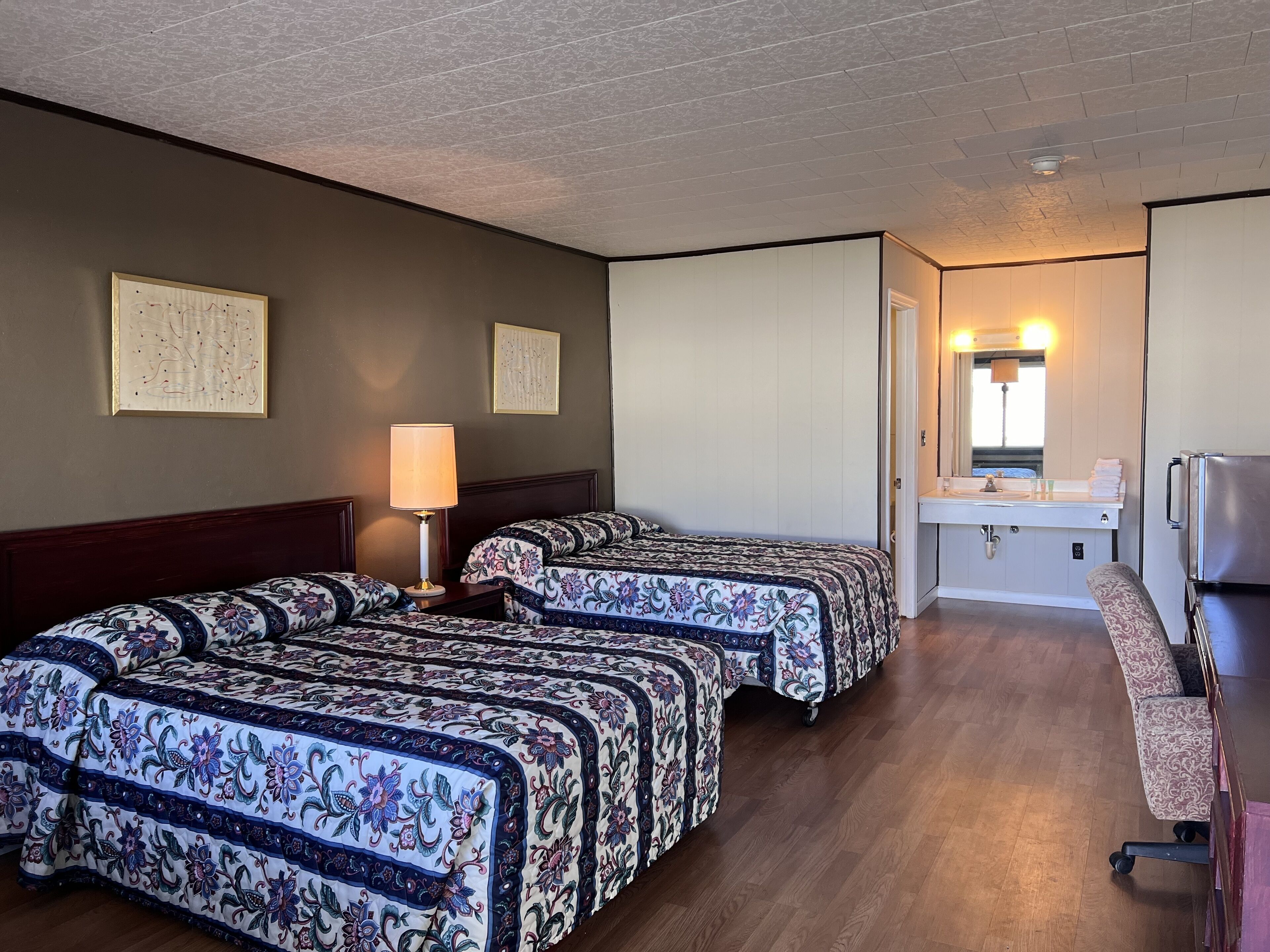Ein Hotelzimmer mit zwei Betten, einem Schreibtisch, einem Stuhl, einem Waschbecken und einem Spiegel.
