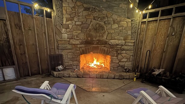 A stone fireplace with a fire, two lounge chairs, and a wooden fence. - Glenwood Inn - Stroudsburg (Delaware Water Gap)