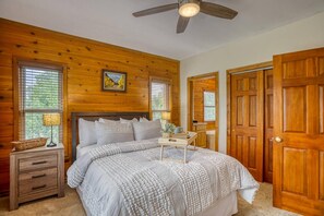Une chambre avec un ventilateur de plafond en bois, un lit recouvert d’une couvre-lit à motifs blancs et gris, une table de chevet avec une lampe et une fenêtre avec des stores.