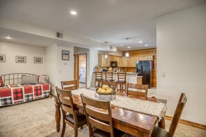 A dining area with a wooden table and chairs, a kitchen in the background, and a bed with a checkered blanket.