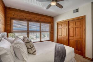 A bedroom with a bed, a ceiling fan, wooden walls, a window with blinds, and a wooden door.