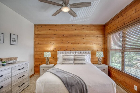 Une chambre avec un mur en bois, un lit avec une literie blanche, deux tables de chevet avec des lampes et un ventilateur de plafond.