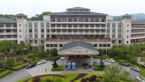 A multi-story hotel with a central entrance, surrounded by landscaped gardens and parked cars. - Huitang Zilongwan Hot Spring Holiday Hotel (Changsha)