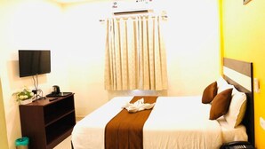 A hotel room with a bed, a TV, a small desk with a plant, a trash bin, and a window with curtains. - Royal Residency (Sriperumbudur)