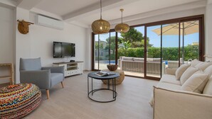 A spacious living room with a grey sofa, a round coffee table, a television, and a large window offering a view of greenery and a pool.