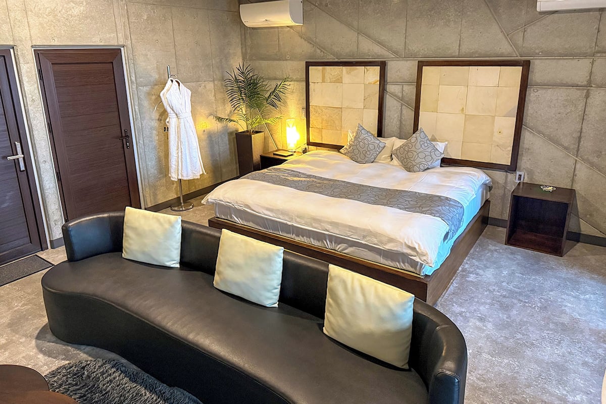A modern bedroom with a large bed, a sitting area with a sofa, a potted plant, and a robe hanging on a hook.