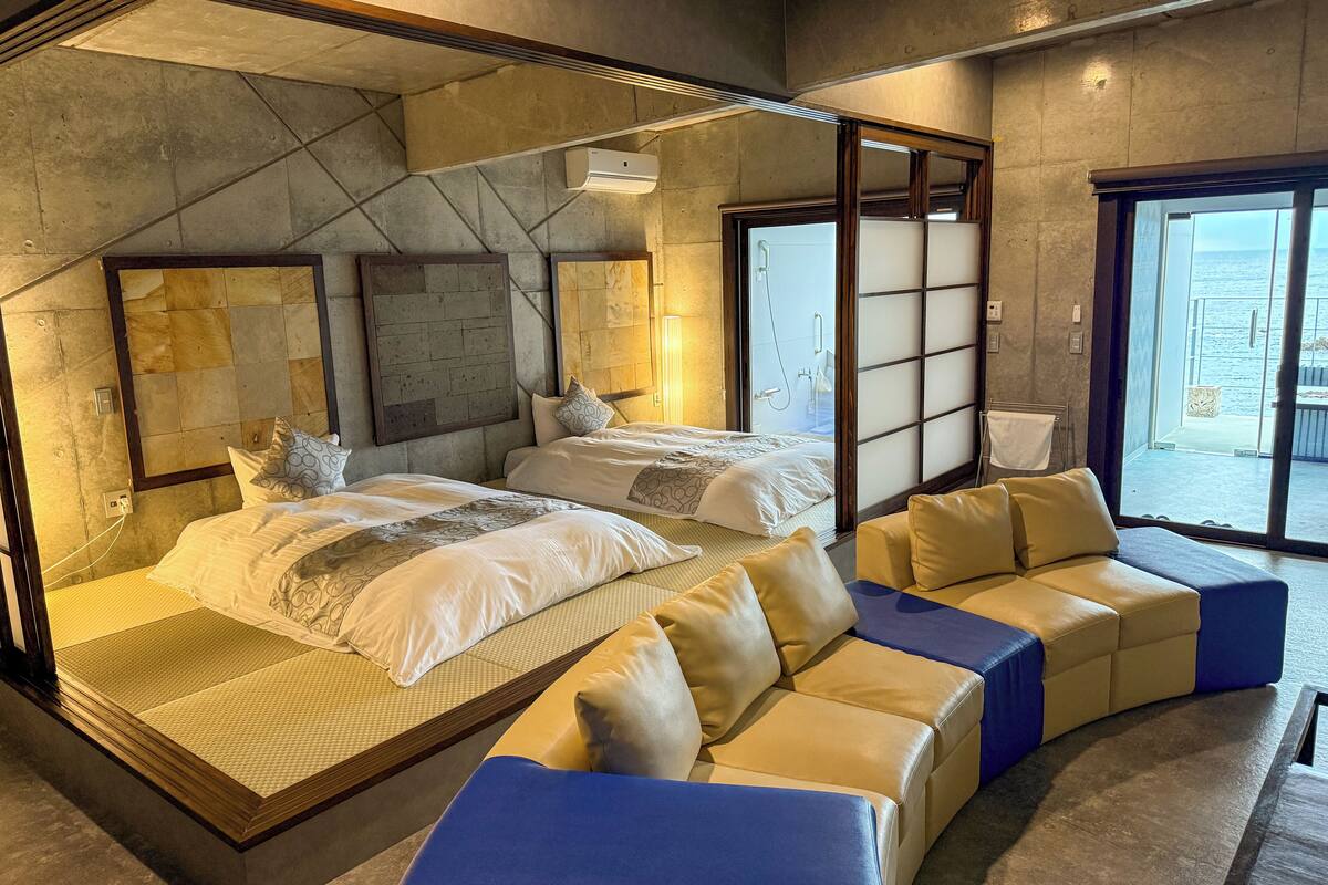 A modern hotel room with a large bed, a sofa, and a sliding glass door leading to a balcony.