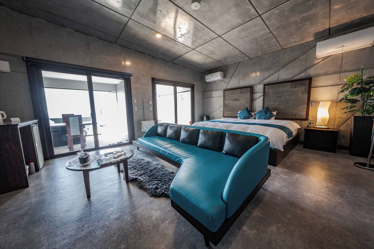 A modern hotel room with a large bed, a sofa, a small table, and a view of the outdoors.
