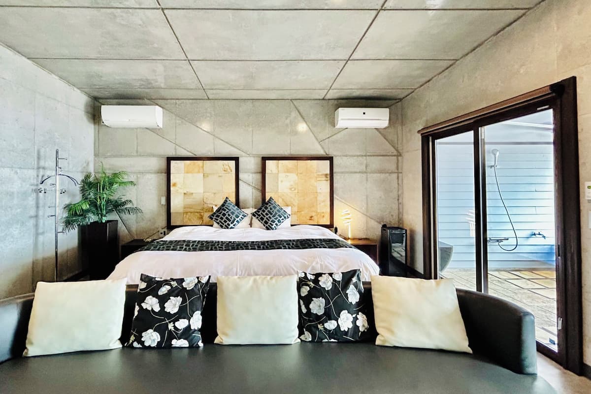 A modern bedroom with a large bed, a potted plant, and a sliding glass door leading to a balcony.