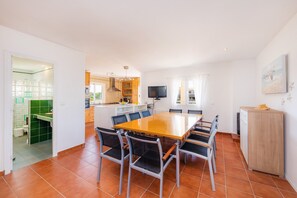 Un comedor espacioso con una gran mesa y sillas, una cocina al fondo y un televisor montado en la pared.