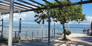 En strandpromenad med en pergola, sittplatser och utsikt över havet och de avlägsna bergen.