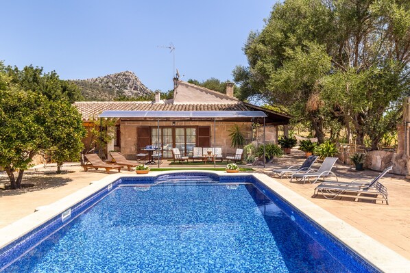 Une piscine bordée de carreaux, un bâtiment avec un toit recouvert de tuiles, et un paysage arboré avec une montagne en arrière-plan.