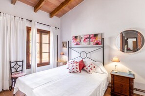 A bedroom with a bed, a nightstand, a chair, a window with curtains, and a wall-mounted mirror.