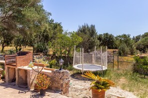 Un espace patio comprenant un barbecue en briques, des plantes en pot, un trampoline et une clôture en métal.