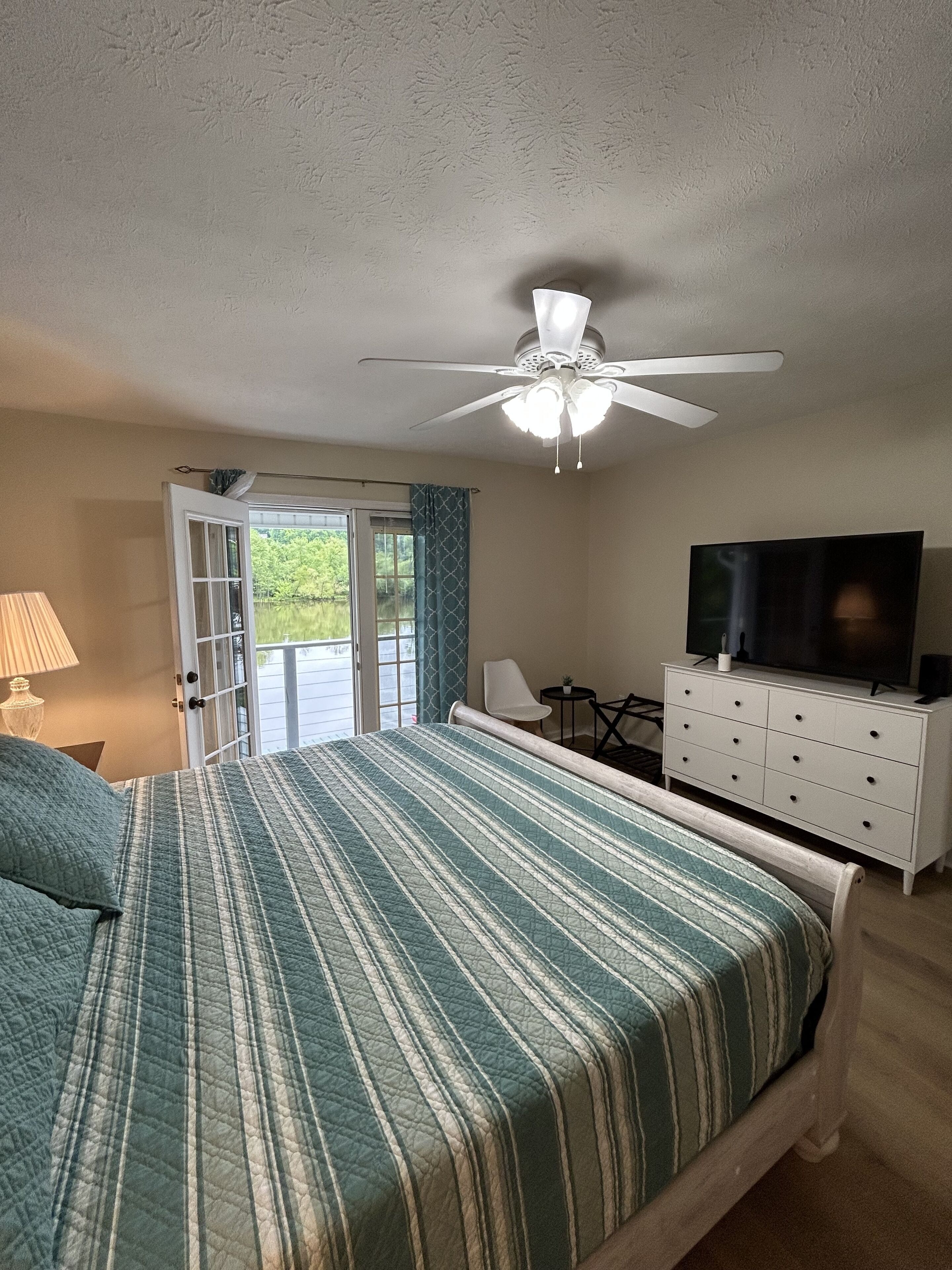 A bedroom with a bed, a ceiling fan, a TV, a desk, and a window with curtains.