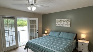 Quarto com cama, ventoinha no teto, mesa de cabeceira, luminária e vista por uma porta.