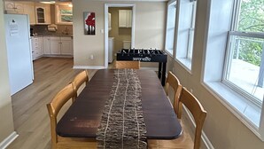 Uma área de refeições com mesa e cadeiras de madeira, cozinha ao fundo e grandes janelas.