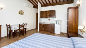 A room with a bed, a dining table with chairs, wooden cabinets, a kitchenette, and a ceiling with exposed beams. - RESIDENZA LA SCOGLIERA - MONO N° 6 LA SCOGLIERA (Rio Marina)