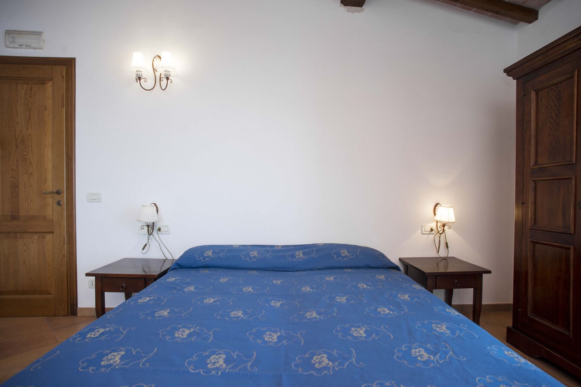 Un dormitorio con una cama, dos mesitas de noche, un armario de madera y lámparas de pared.