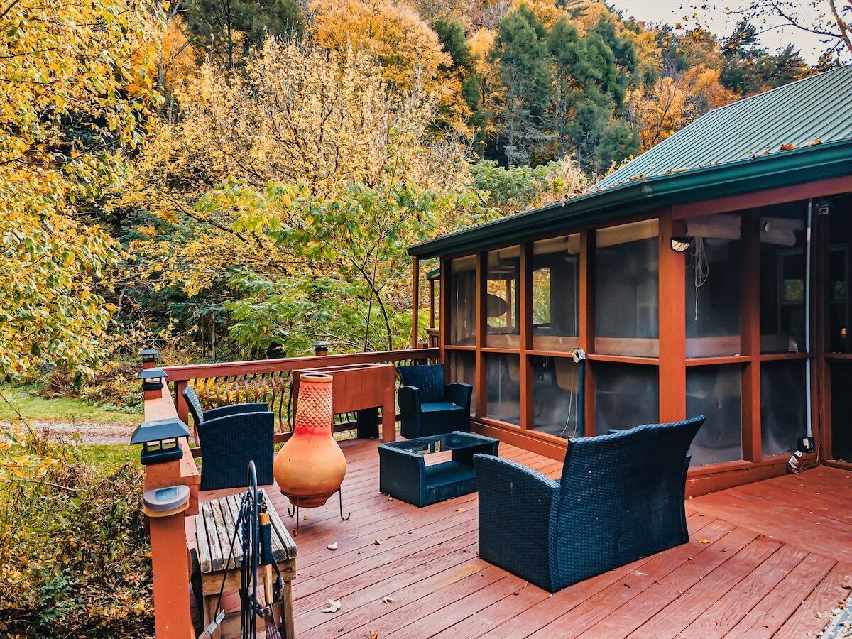A wooden deck with outdoor furniture, a glass-enclosed room, and a view of autumn foliage.