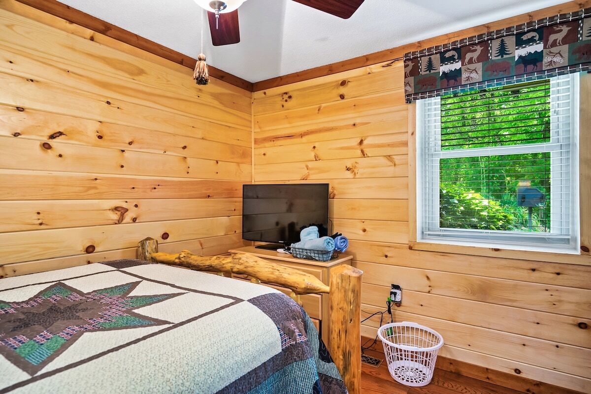 A wooden cabin room with a bed, a television, a window with a view of greenery, and a wooden beam.