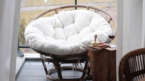 Una silla de mimbre con un cojín blanco, una mesita de madera con una botella y vasos, y vistas a través de un ventanal.