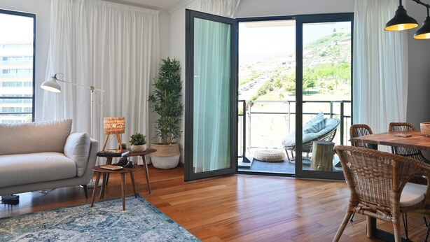 Una sala de estar moderna con un sofá, una mesita de noche con una lámpara, una planta en maceta y un comedor con sillas de mimbre.