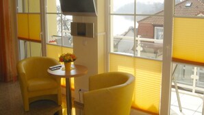 Una stanza con due poltrone gialle, un tavolino con un vaso di fiori, una televisione a schermo piatto montata a parete e ampie finestre che offrono vista su una casa e montagne.