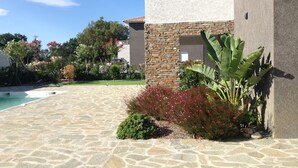 Uma piscina de pedra com céu azul límpido, um muro de pedra com janela e vegetação exuberante, incluindo uma bananeira.