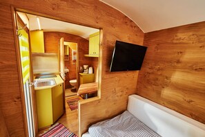 Una camera compatta, con rivestimenti in legno, un letto, un angolo cottura piccolo e una TV fissata al muro.