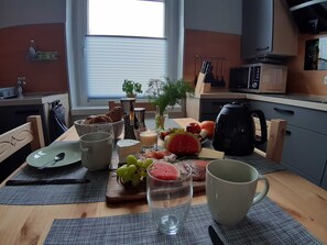 A breakfast setup with a wooden table, a cutting board with fruits and cheese, a candle, and a kettle.