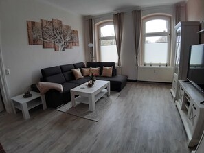 Ein Wohnzimmer mit schwarzem Sofa, weißem Couchtisch und einem Flachbildfernseher auf einem Standfuß.