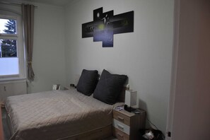 A bedroom with a bed, two pillows, a bedside table with a lamp, a wall art piece, and a window with curtains.