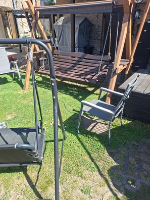 Ein Holzschaukel, eine Metallbank und ein überdachter Sitzbereich in einem Garten mit einer Ziegelmauer und einem Metalltor.