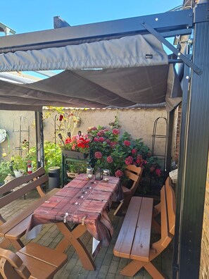 Une terrasse extérieure avec une table et des bancs, entourée de plantes et de fleurs en pot.