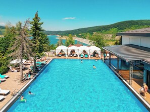 A swimming pool with clear blue water, surrounded by lounge chairs and umbrellas, with a view of a forested hillside and a body of water in the background. - Radika Rezort (Leunovo)